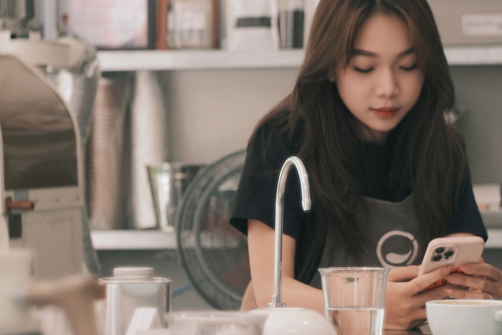 Barista sau quầy nhìn điện thoại, khách ngồi phía sau quán